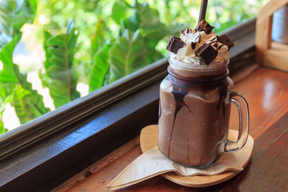 chocolate milkshake in glass topped with whipped cream and chocolate chunks placed on a windowsill 